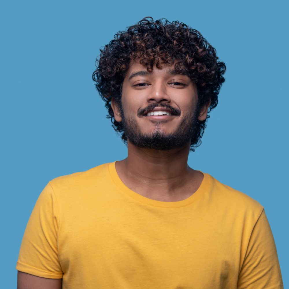 Cute dark-haired guy with a happy expression looking ahead Portrait of a smiling pleased man with curly hair posing for the camera against the blue background